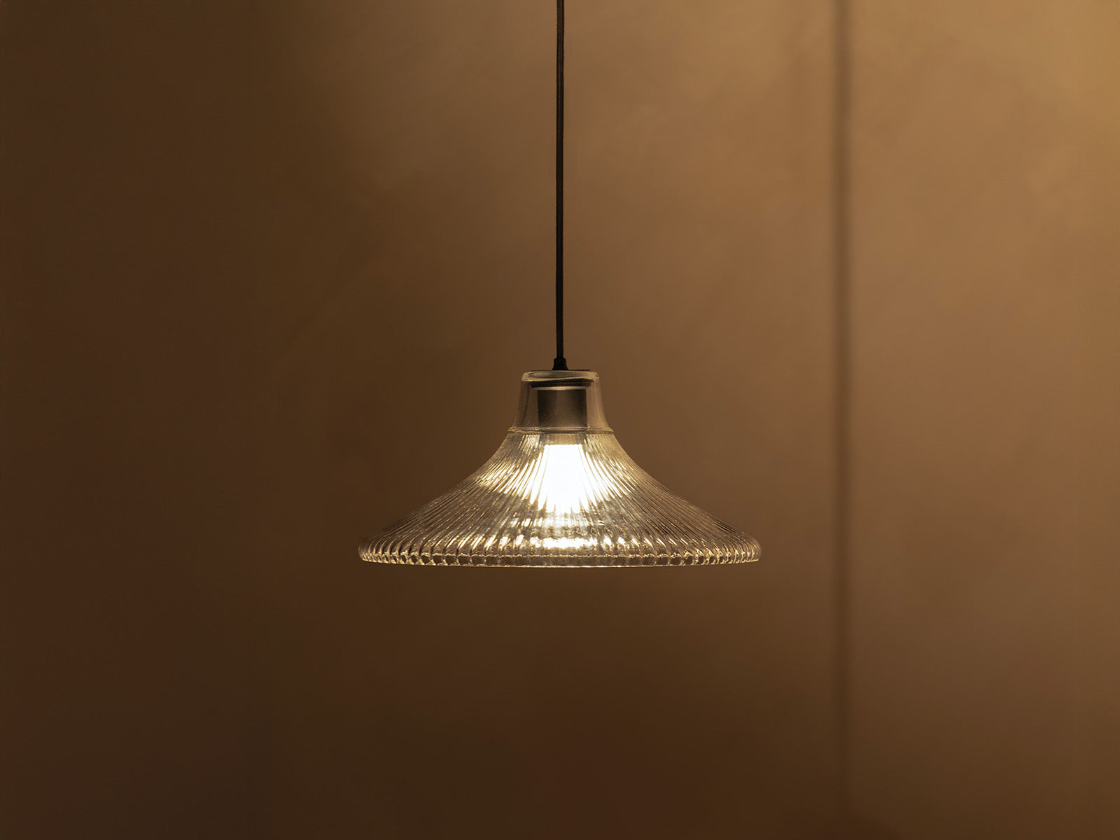 A modern pendant lamp with a glass shade, casting warm light against a muted brown wall background.