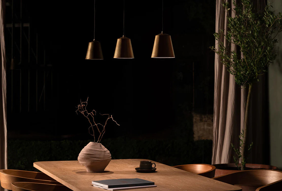 A cozy dining space with a wooden table, minimal decor, three pendant lights, a sculptural vase, and a coffee cup, surrounded by soft shadows.
