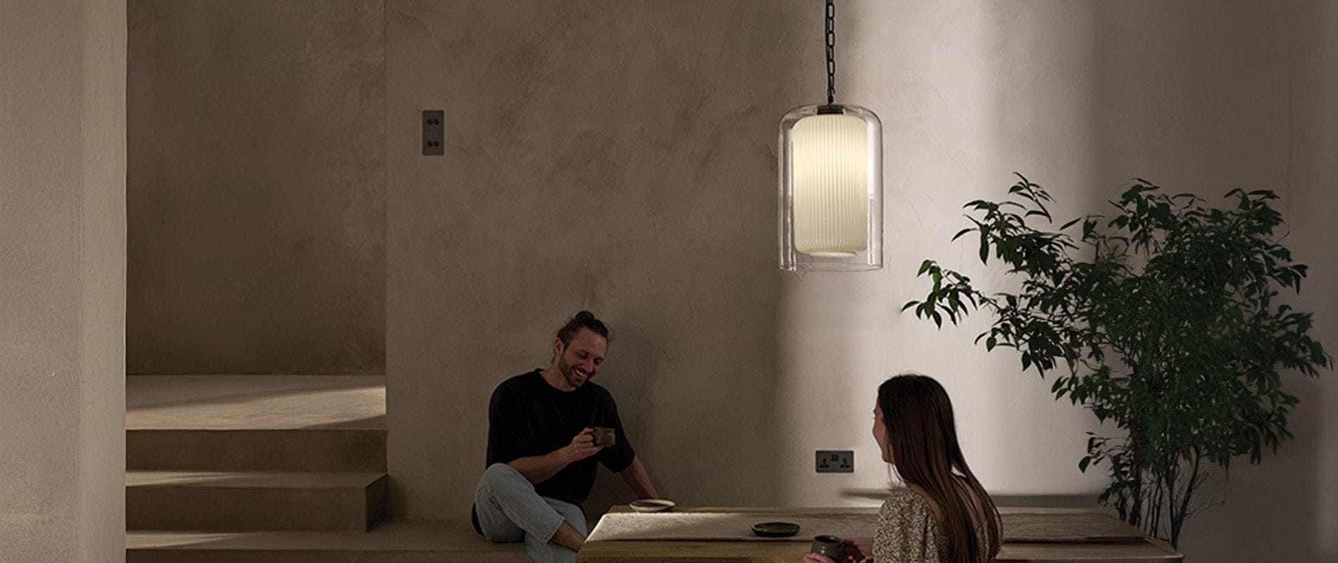 A cozy interior with textured walls, a pendant lamp, and a wooden table, where two people enjoy drinks in a tranquil atmosphere.