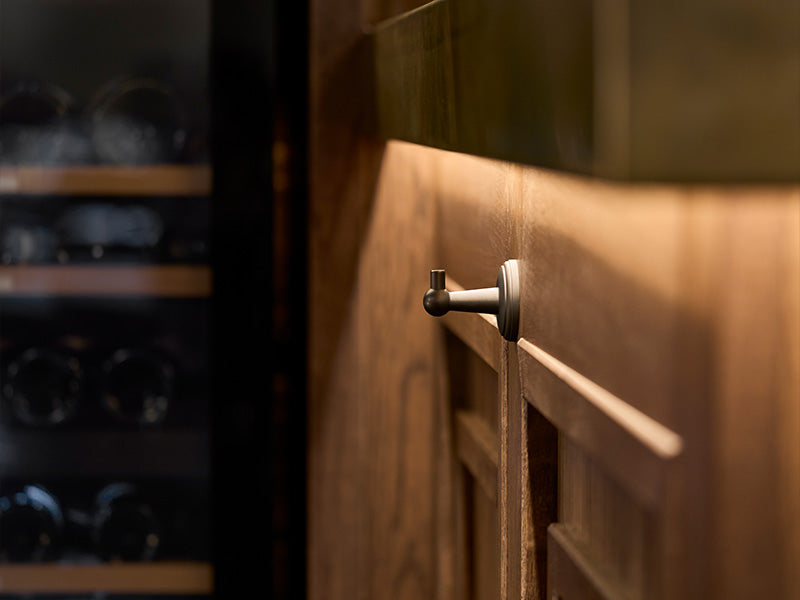 Close-up of a modern, sleek cabinet door handle on a wooden cabinet, with a softly lit background showcasing a wine rack.