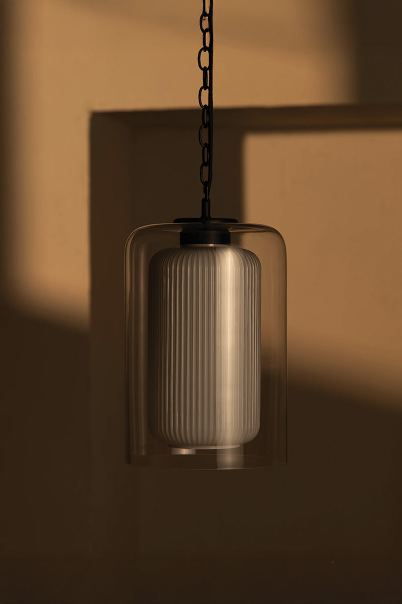 A stylish pendant light features a ribbed white bulb within a transparent shade, suspended by a black chain against a warm-toned wall.