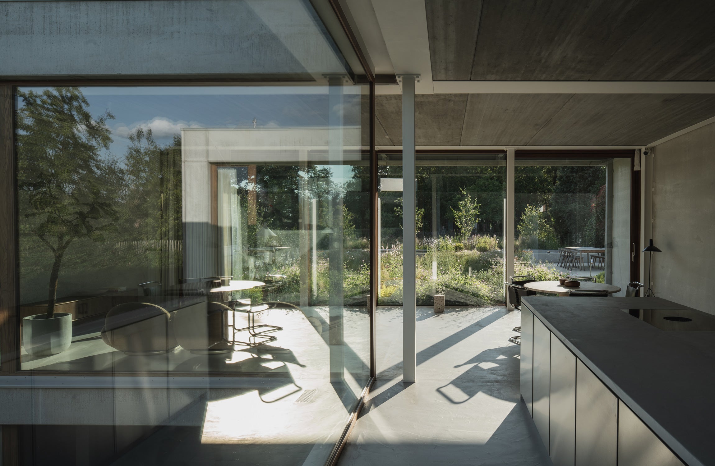 Interior view of a modern kitchen with large glass windows, showcasing a garden outside and minimalist furniture. Sunlight casts shadows inside.