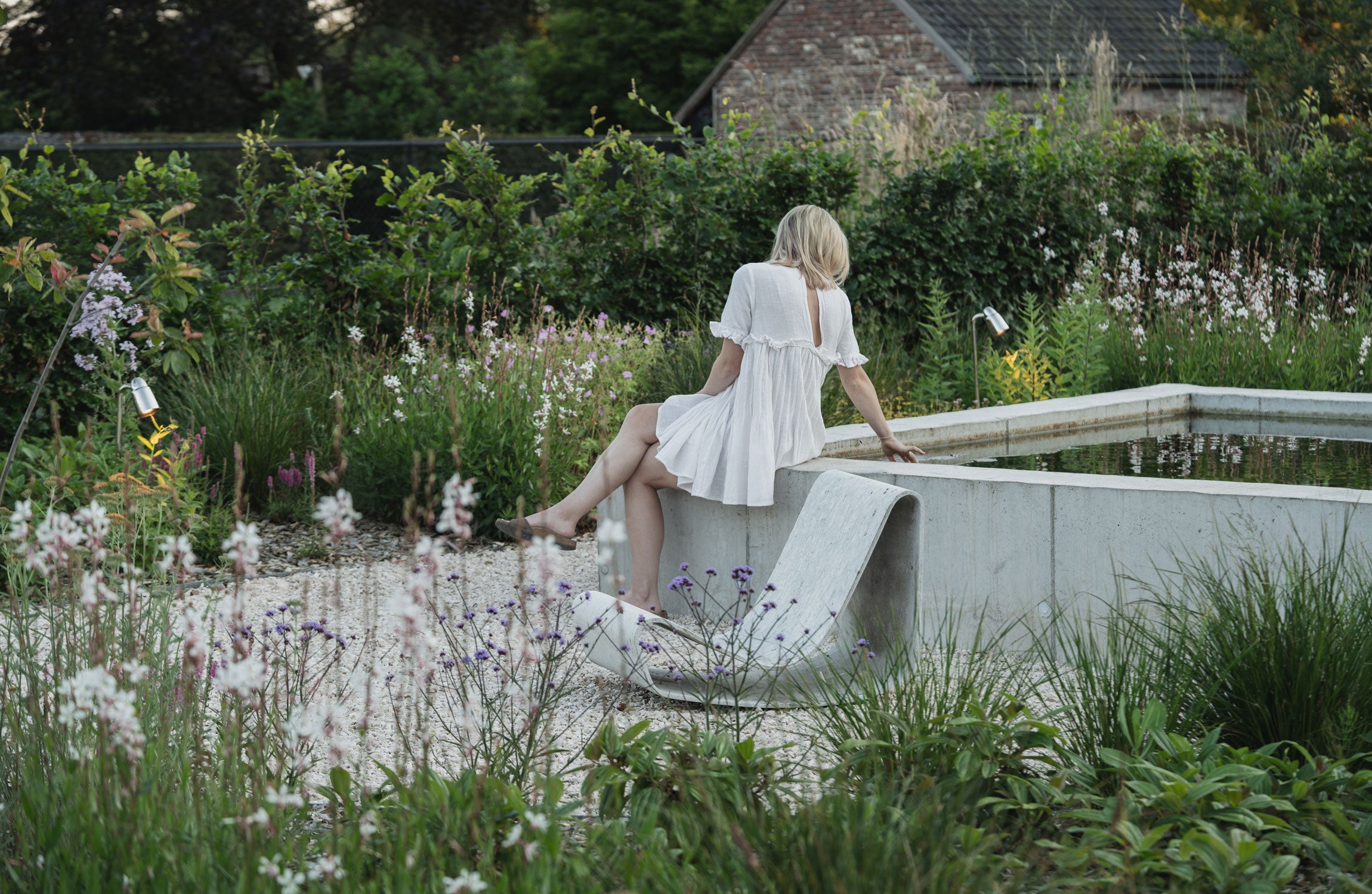 A woman in a white dress sits gracefully on a concrete bench, surrounded by colorful flowers and greenery in a serene garden.
