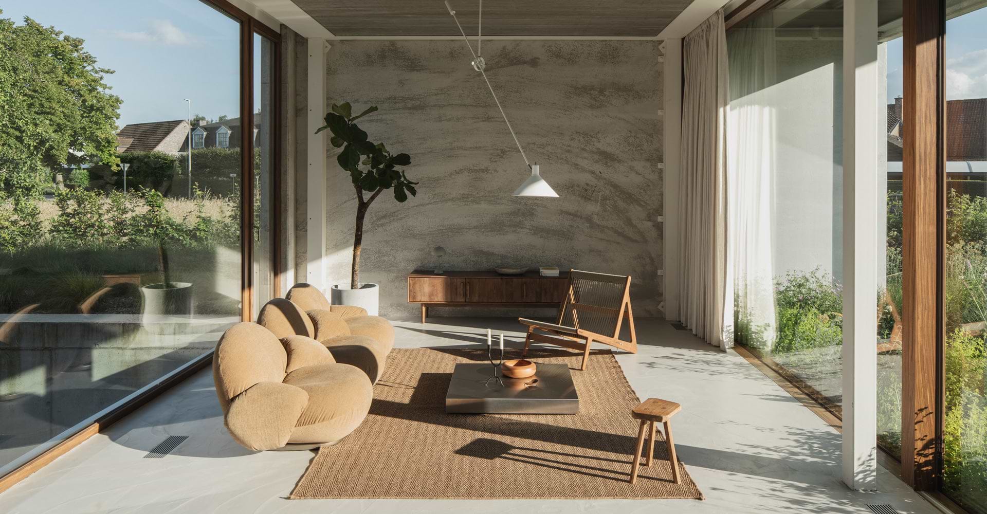 A modern, sunlit living room features curved beige sofas, a wooden coffee table, and a potted plant, creating a cozy atmosphere.