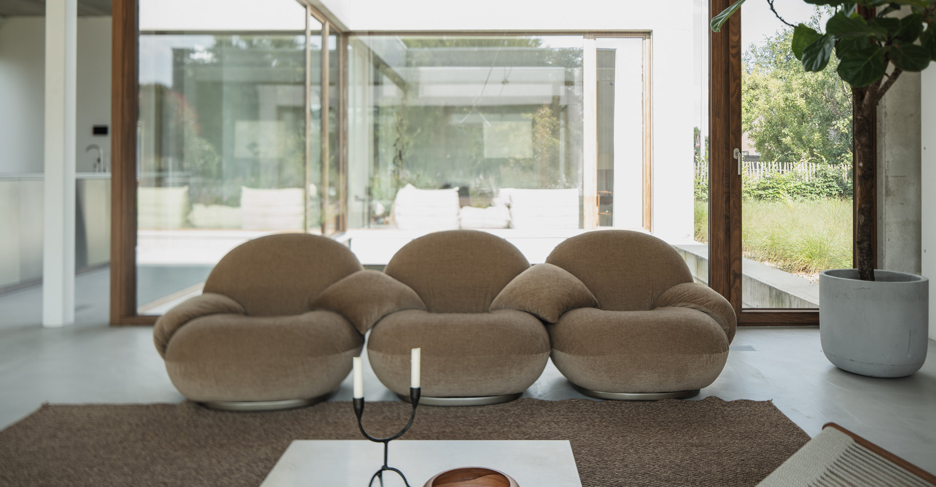 A cozy, rounded beige sofa set in a bright, modern living space with large glass windows showcasing lush greenery outside.