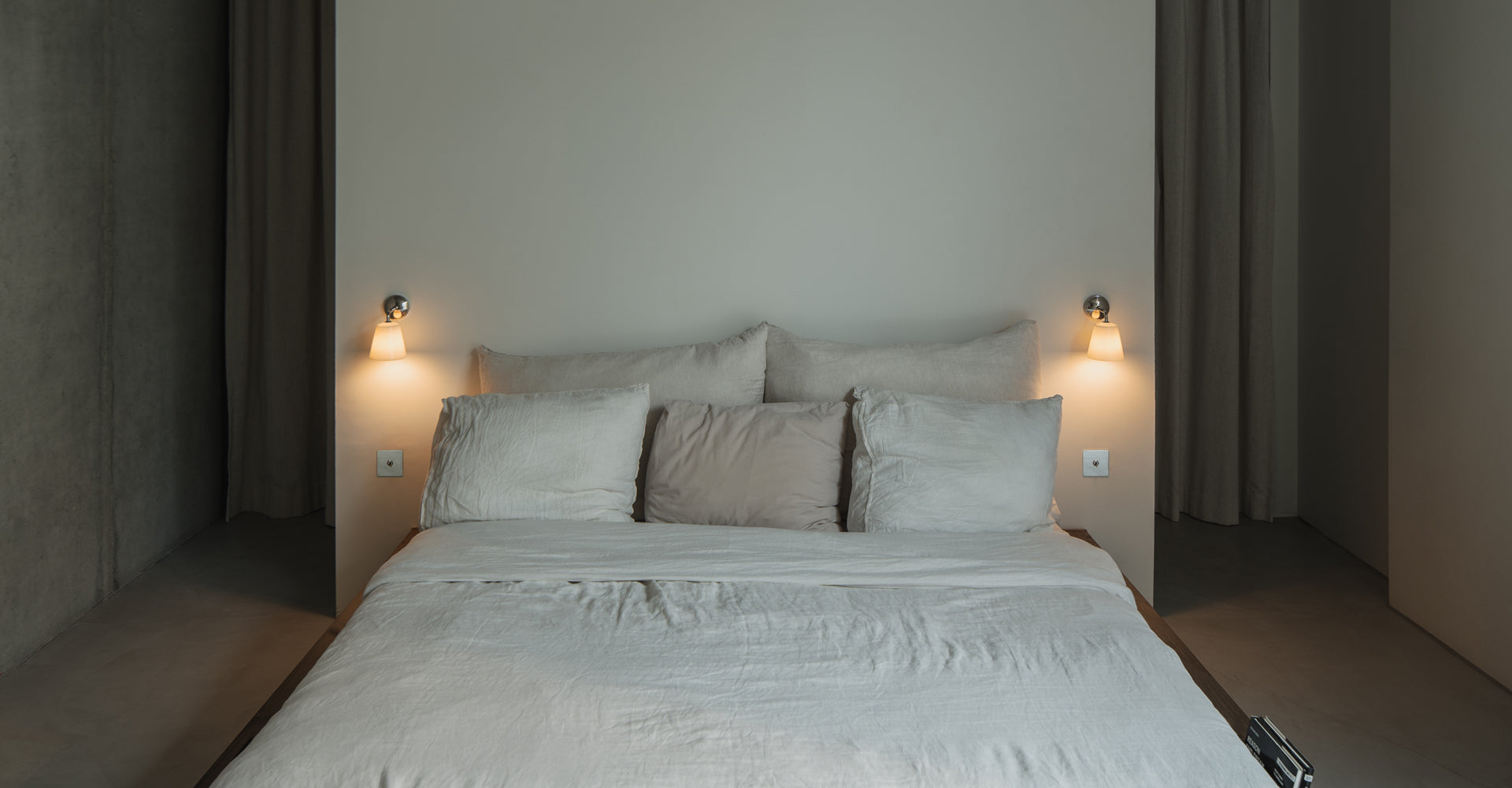A minimalist bedroom featuring a soft, unmade bed with light beige linens and pillows, illuminated by stylish wall sconces.