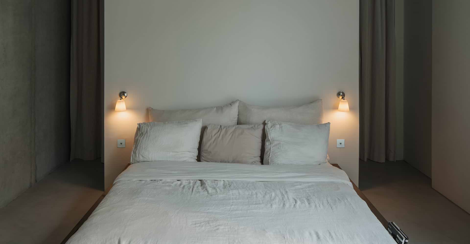 A minimalist bedroom featuring a soft, unmade bed with light beige linens and pillows, illuminated by stylish wall sconces.