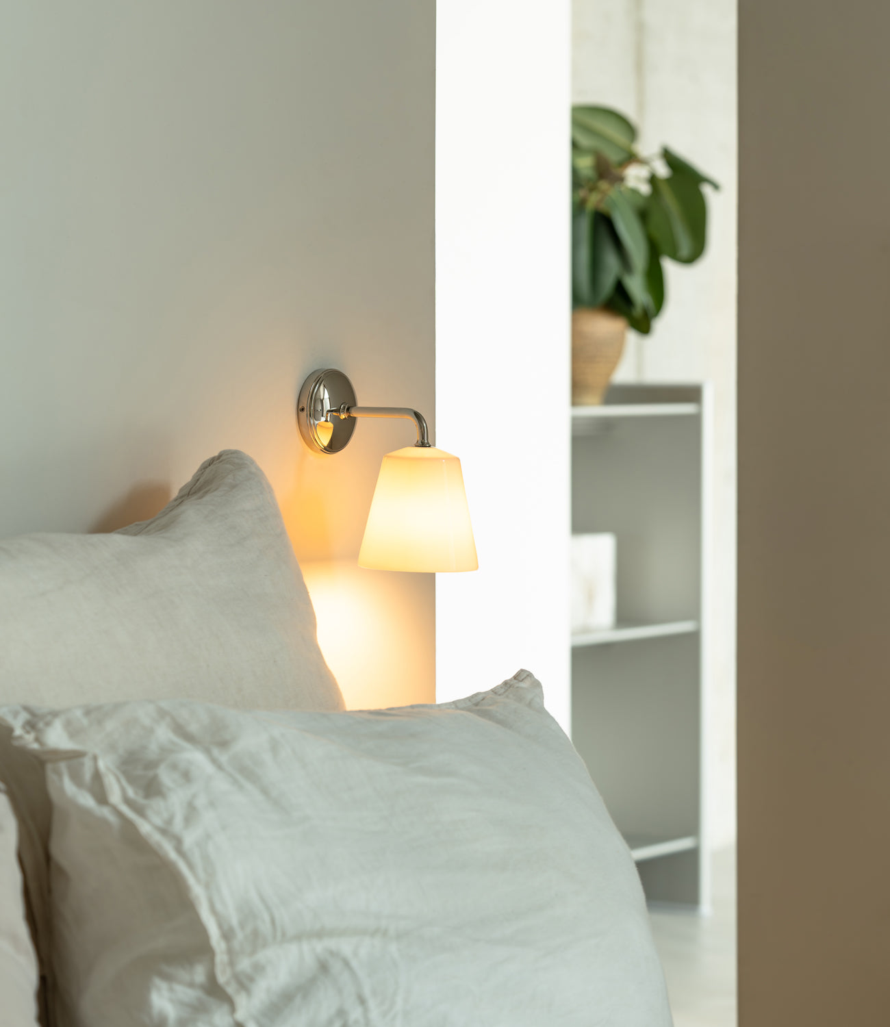 A cozy bed with white linens and a softly glowing wall lamp, set in a modern room with greenery and minimal decor.