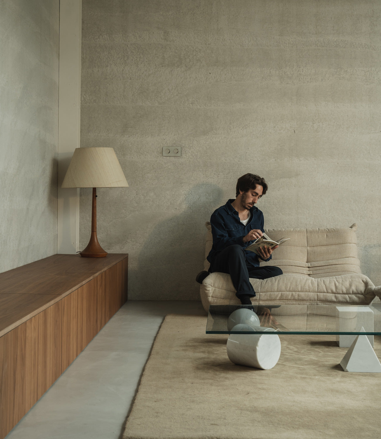 A person seated on a beige couch, reading a book, with a modern glass coffee table, wooden sideboard, and decorative lamp nearby.
