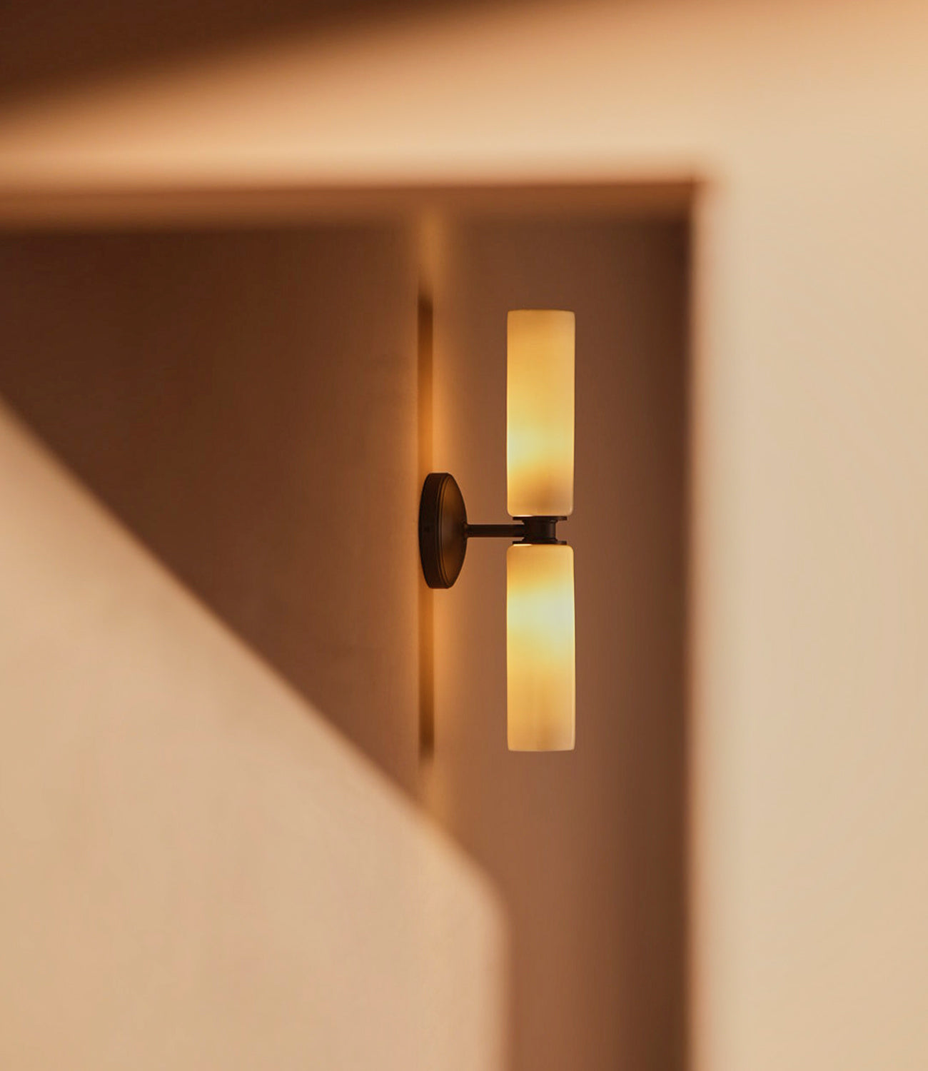 A modern wall sconce with two vertical frosted glass shades, mounted on a dark circular base, softly illuminating a warm-toned wall.
