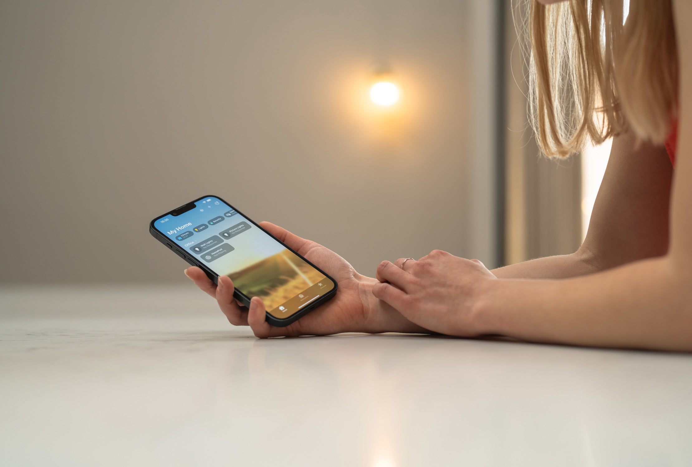 A hand holding a smartphone displaying a home automation app interface with controls for lights and devices.