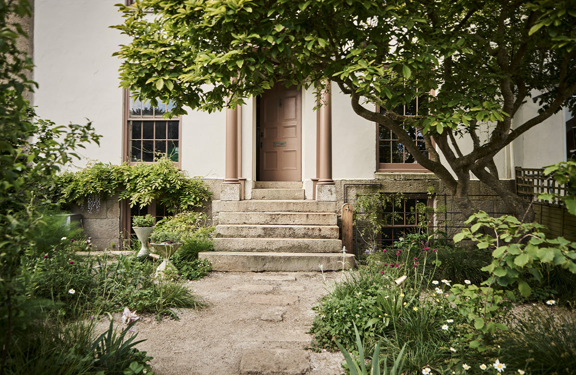 A welcoming entrance with a brown door and stone steps, framed by lush greenery and colourful flowers in a serene garden setting.