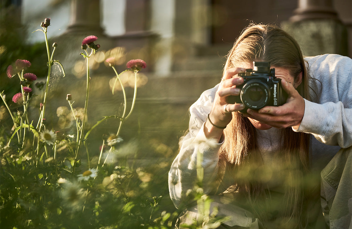 A young person with long hair crouches in a garden, focusing a camera on vibrant flowers in the golden hour light.