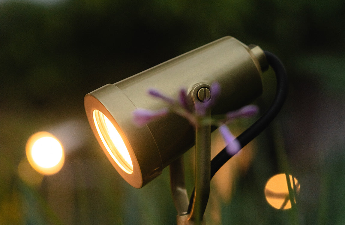 A close-up of a brass outdoor spotlight illuminating purple flowers, with a blurred orb of light in the background at dusk.