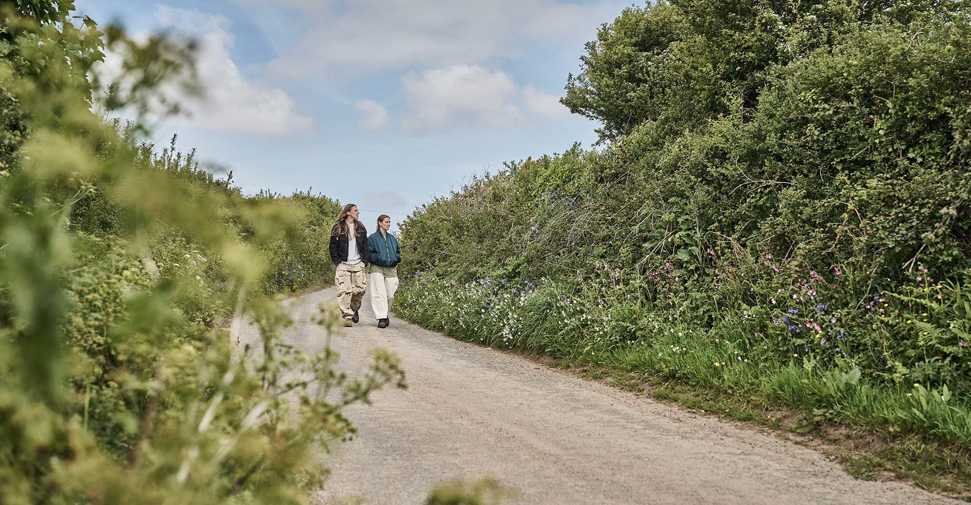 Two people walk along a scenic dirt path lined with lush greenery and wildflowers under a partly cloudy sky.