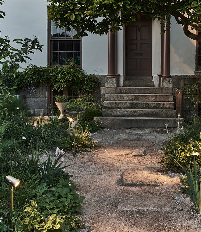 A charming entryway with stone steps framed by lush greenery and soft garden lights illuminating the path to a welcoming door.