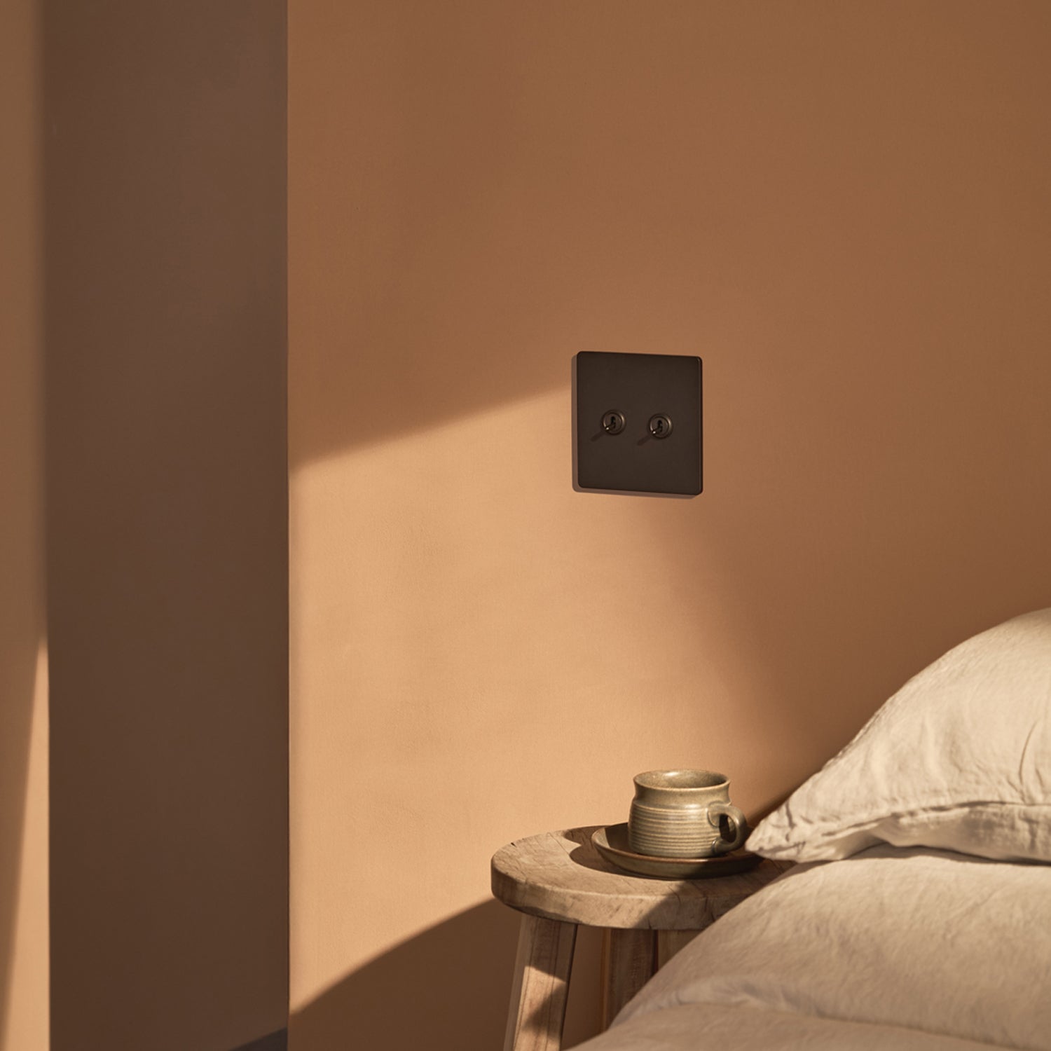 A modern black power outlet on a soft beige wall, with a wooden stool beside a plush bed and a cup resting on the stool.
