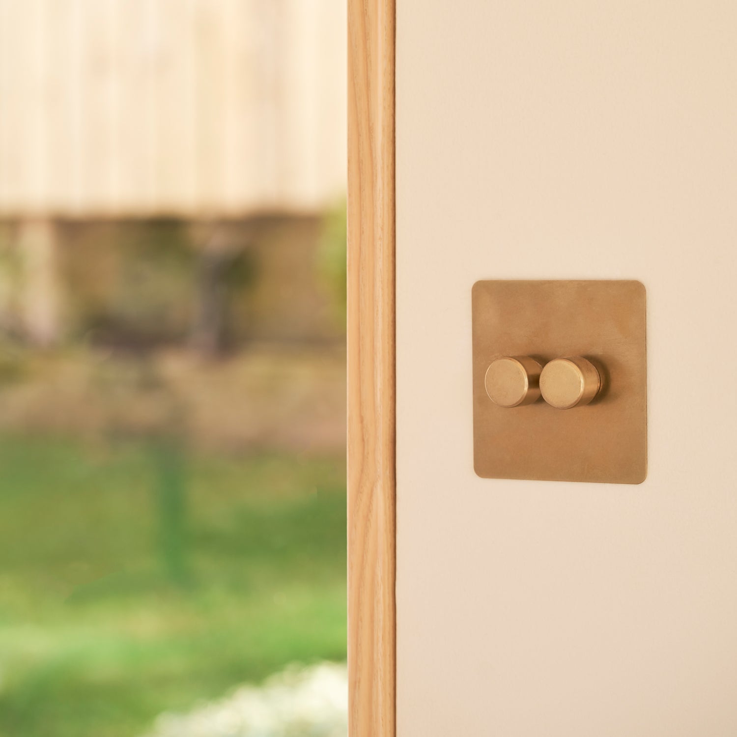 Close-up of a stylish brass light switch mounted on a cream wall, with a blurred garden background featuring greenery and flowers.