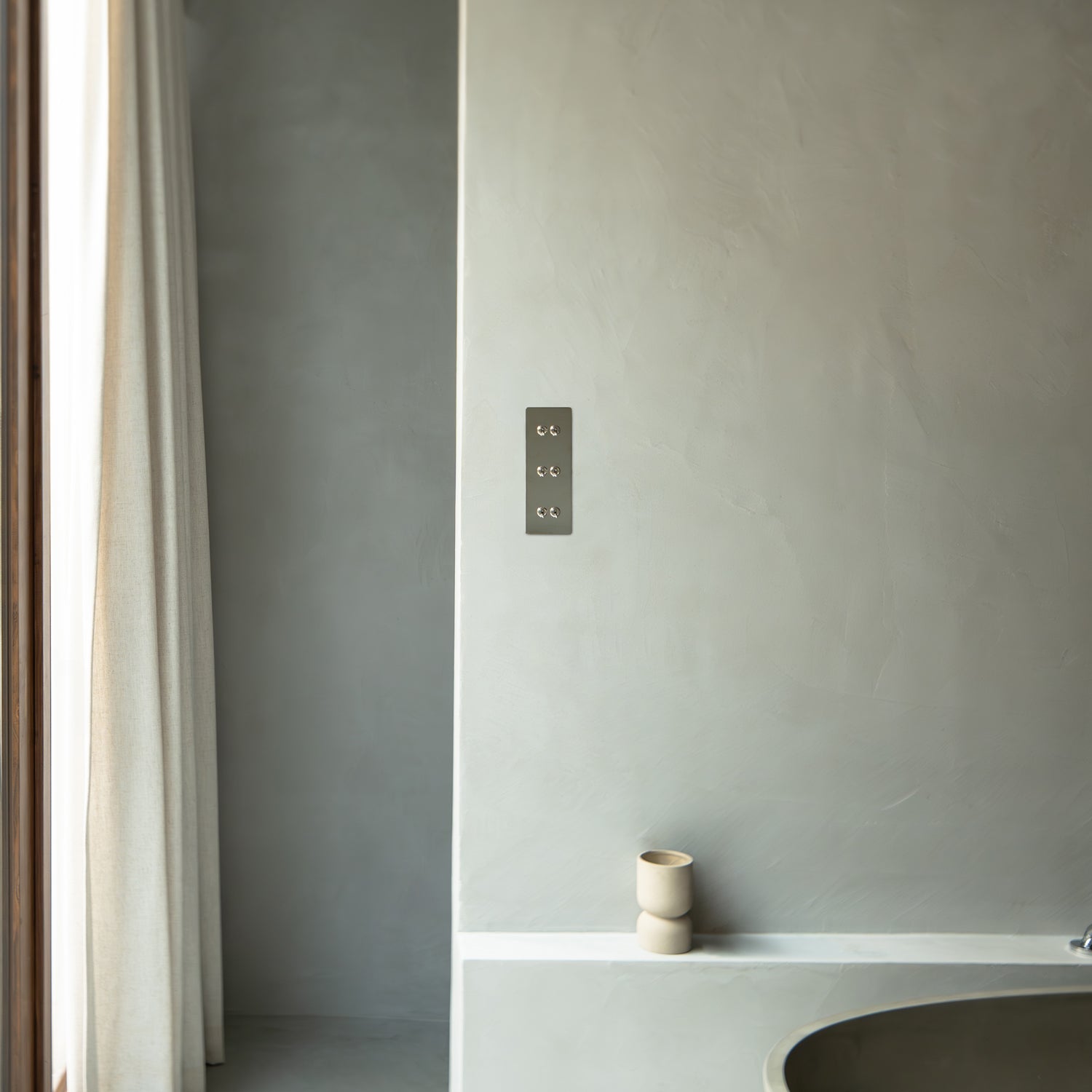A modern bathroom with gray plaster walls, a minimalist tub, and soft curtains allowing natural light to enter.