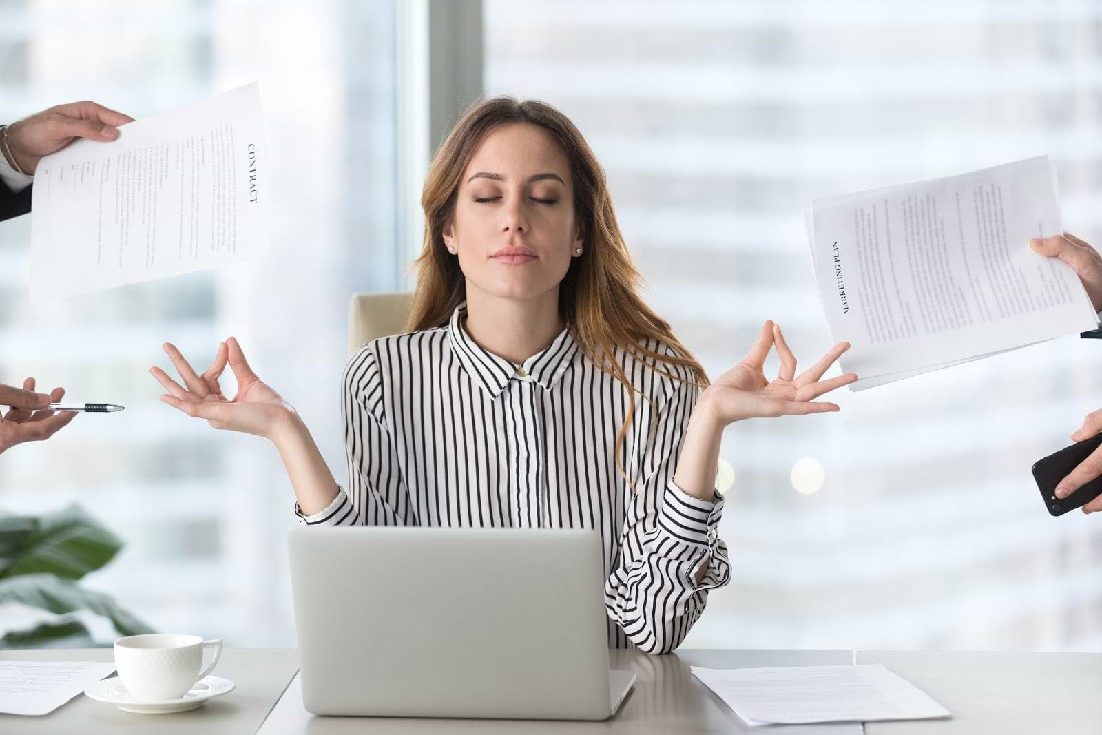Woman meditating with eyes closed in front of laptop as people hold papers in front of her