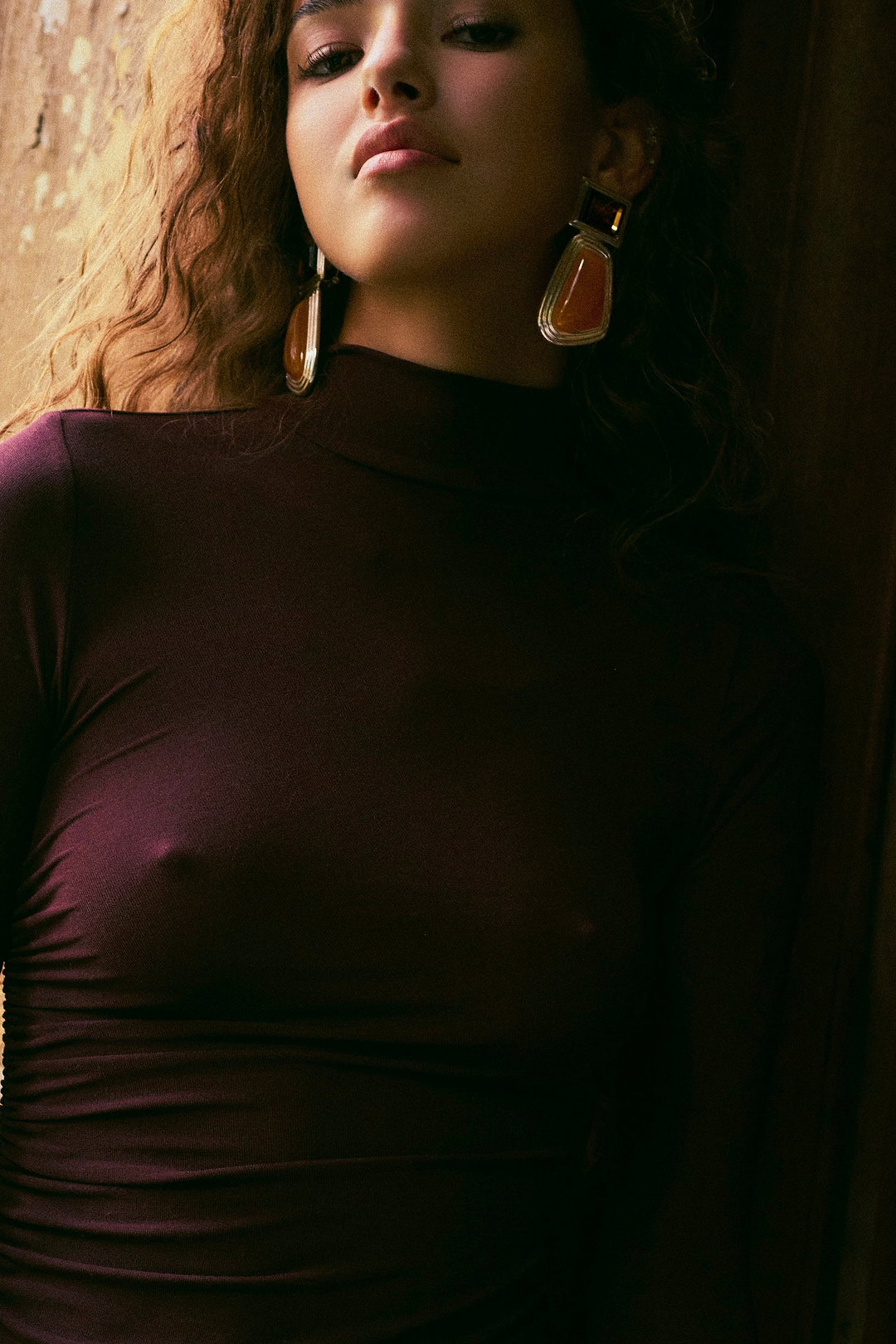 A woman wearing a dark plum long-sleeve mock-neck top with a side cutout, styled with large gold and orange statement earrings, leaning against a dimly lit wooden background.