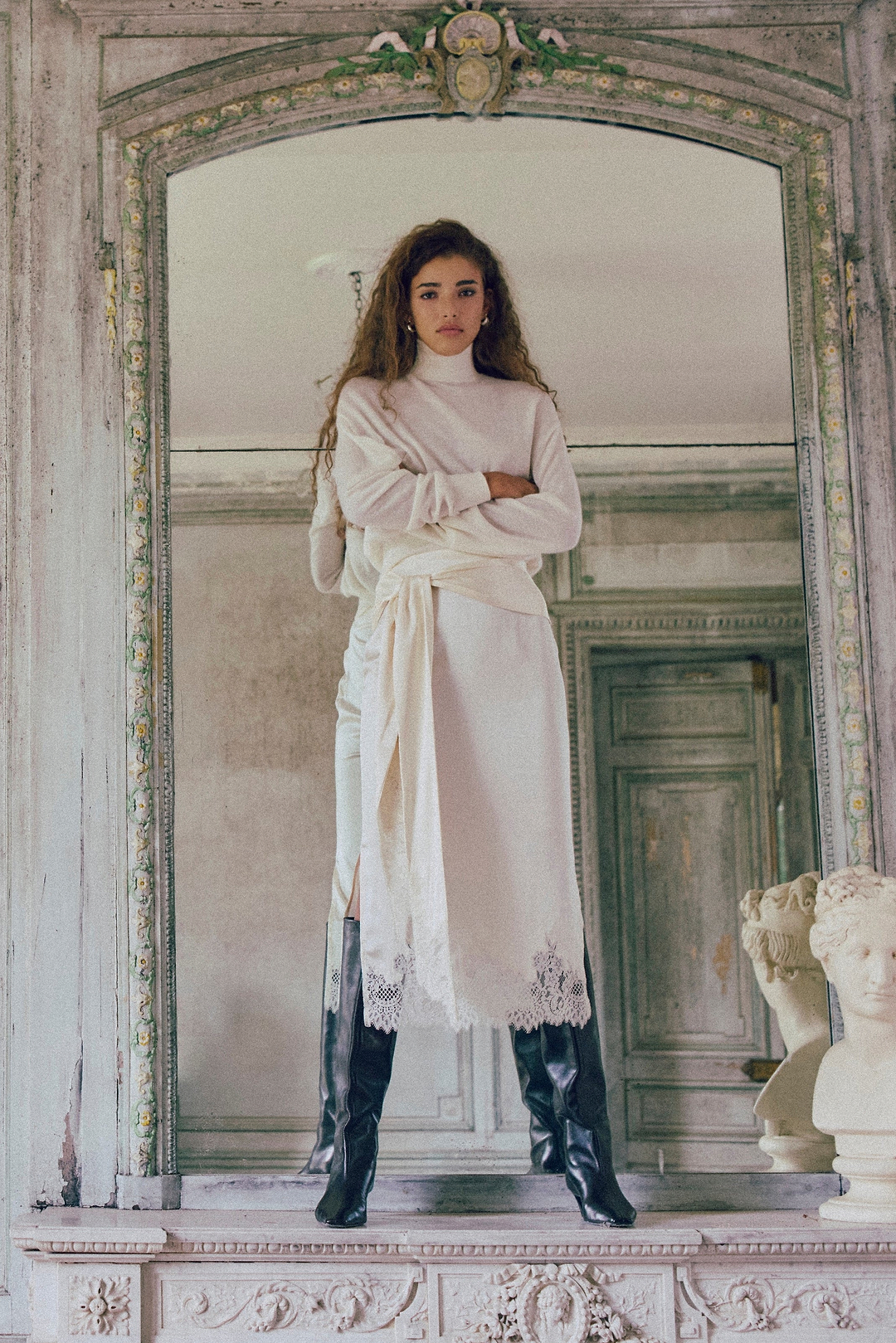 A woman stands in a large ornate mirror within an old stone room, wearing a long white robe with lace trim and black knee-high boots. The background features distressed gray-green doors and classical busts.