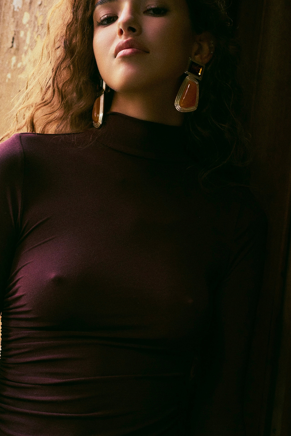 A woman wearing a dark plum long-sleeve mock-neck top with a side cutout, styled with large gold and orange statement earrings, leaning against a dimly lit wooden background.