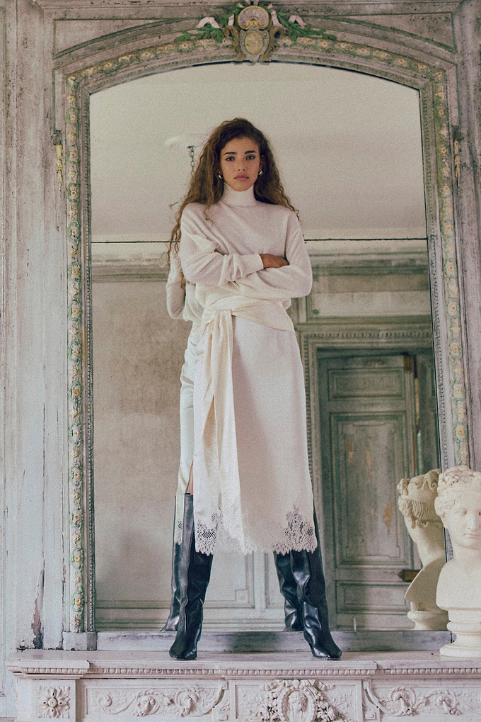 A woman stands in a large ornate mirror within an old stone room, wearing a long white robe with lace trim and black knee-high boots. The background features distressed gray-green doors and classical busts.