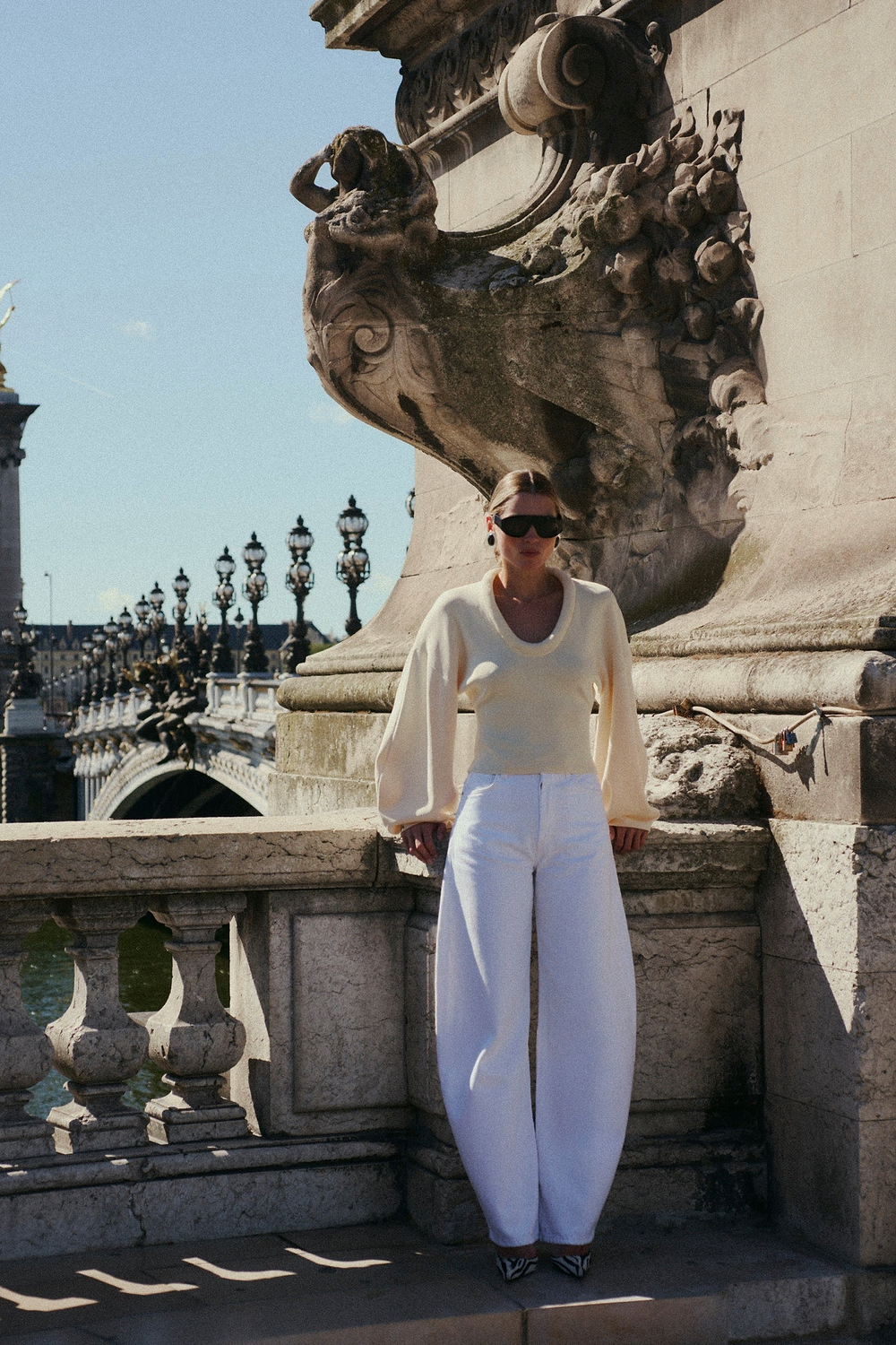 A woman stands on a stone bridge in Paris wearing a cream long-sleeve knit top and white wide-leg trousers, with a beige cap and sunglasses. The background features the ornate stone architecture of the bridge and a distant view of Pont Neuf under a clear blue sky.