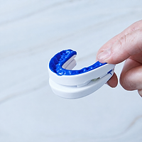 A close-up of a hand holding a white and blue mouthguard against a light gray marble background.