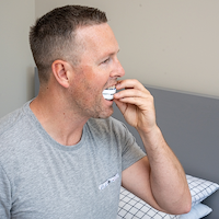 Man in a gray t-shirt using a mouthguard in a bedroom setting