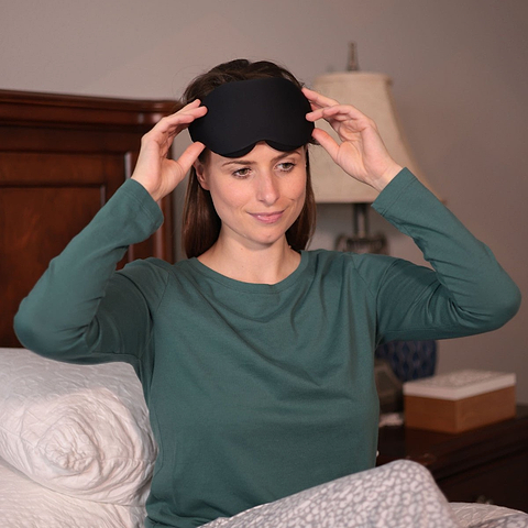 Contoured eye mask for sleeping, worn by a woman in a green shirt, sitting on a bed with pillows and a lamp in the backgro...