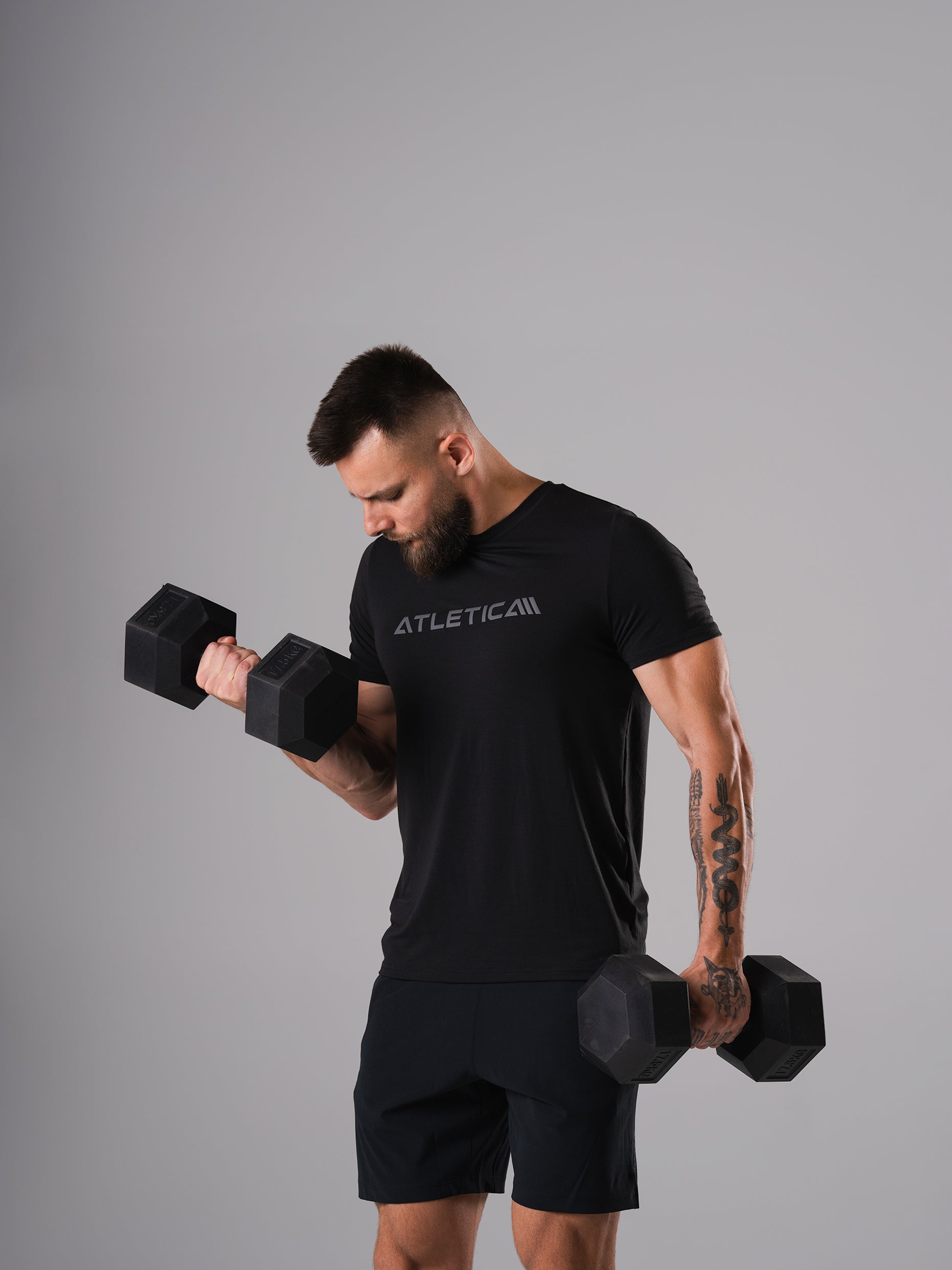 Mann im schwarzen Merch Flex Train T-Shirt von Atletica beim Bizepscurl mit zwei Hexagon Kurzhanteln