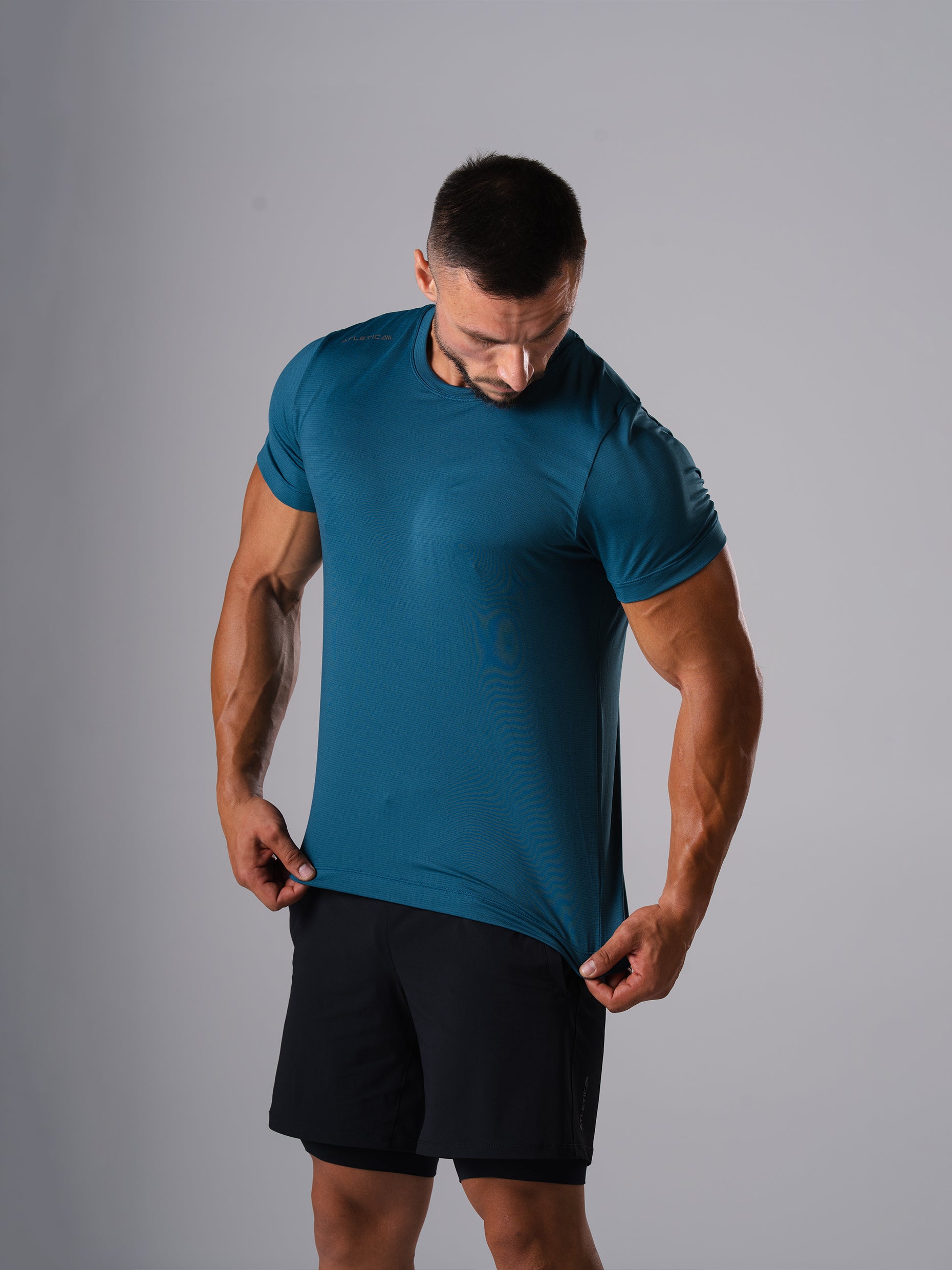 Mann zieht am Saum des Hard Lift T-Shirts in Legion Blue von Atletica, Detailansicht von Stoff und Passform