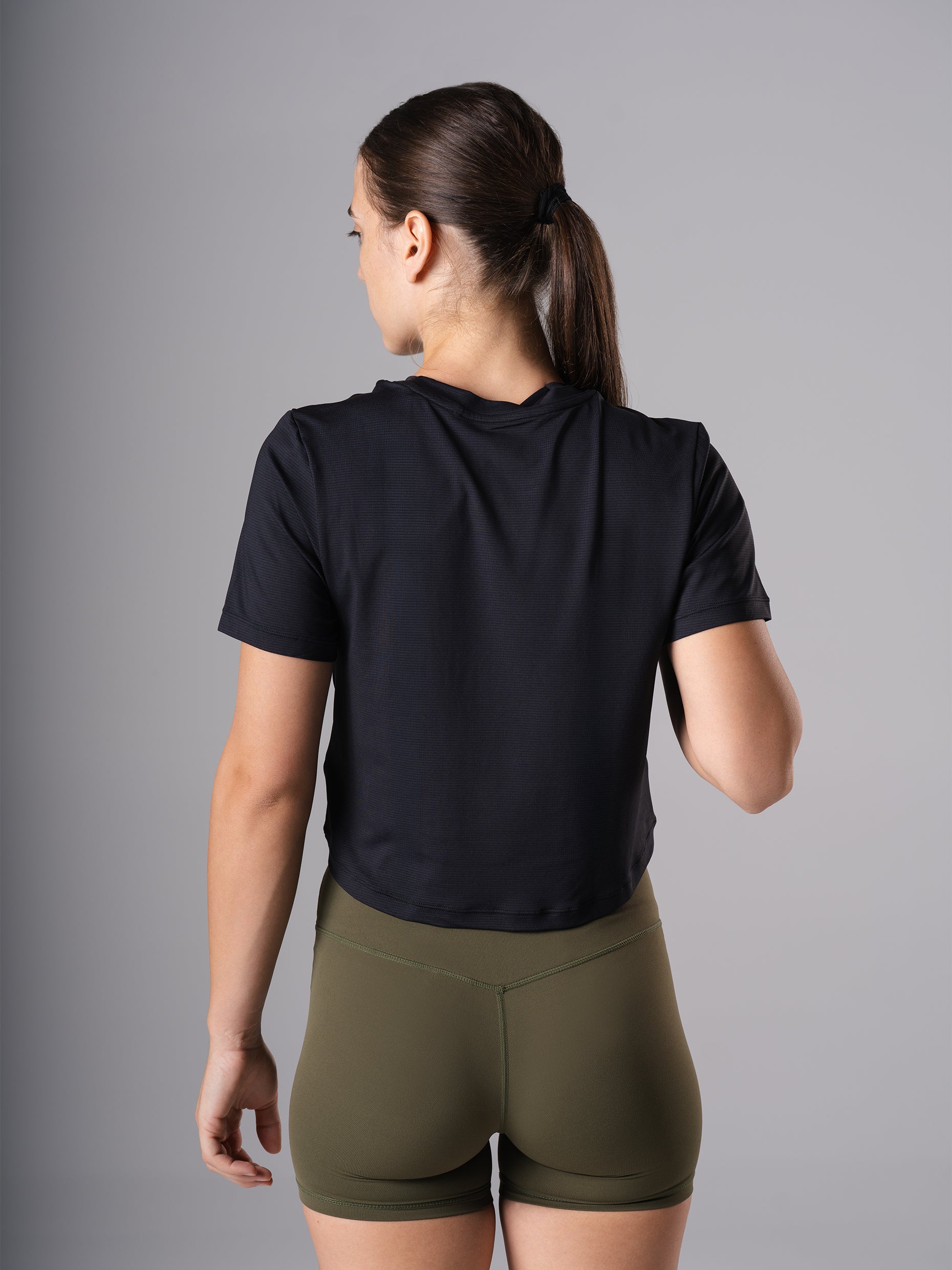 Schräge Rückansicht des Power Lift Cropped T-Shirts in Schwarz von Atletica mit olivgrünen Shorts