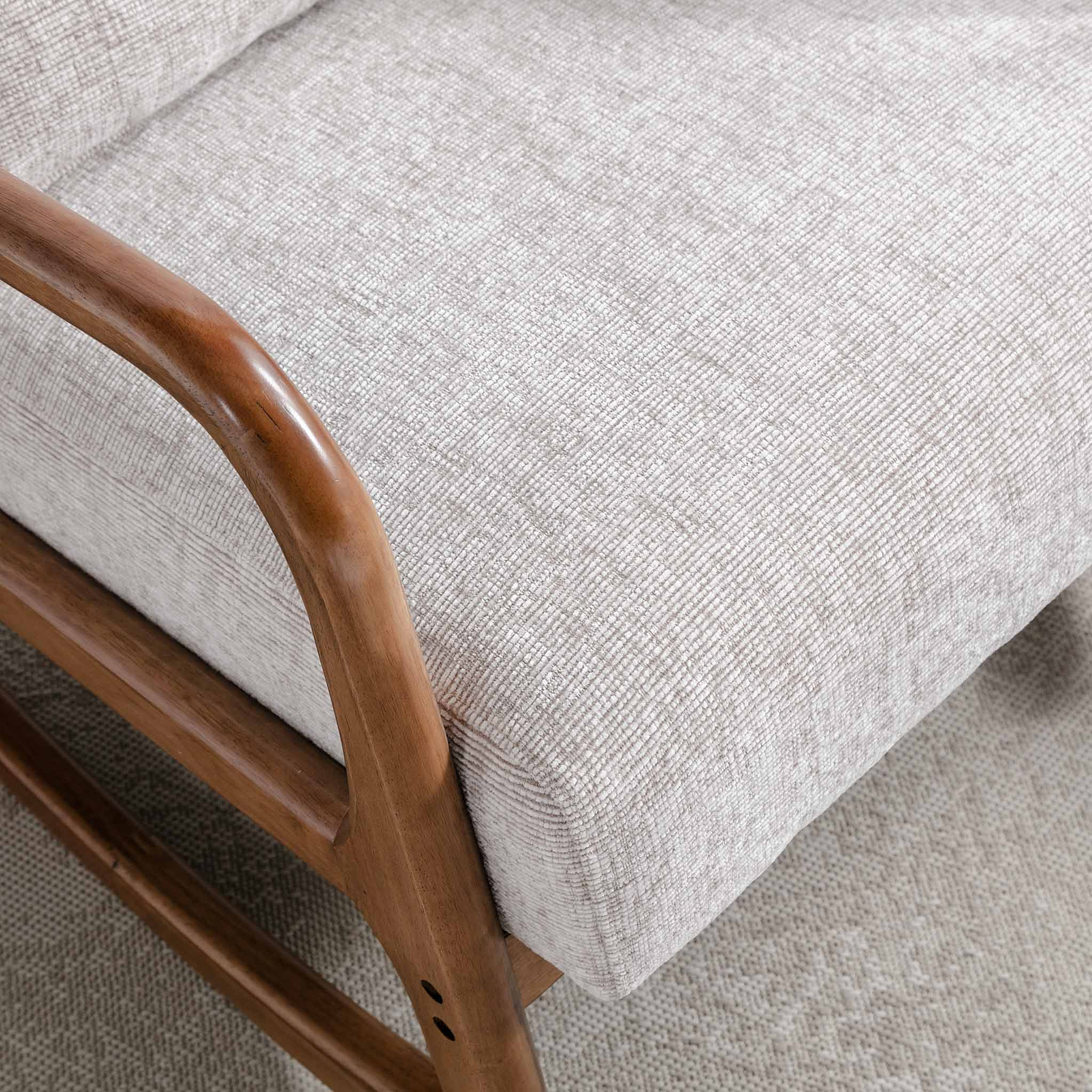 Close-up of a wooden armrest connected to a textured light-grey upholstered cushion, part of a piece of furniture, against a beige carpet.