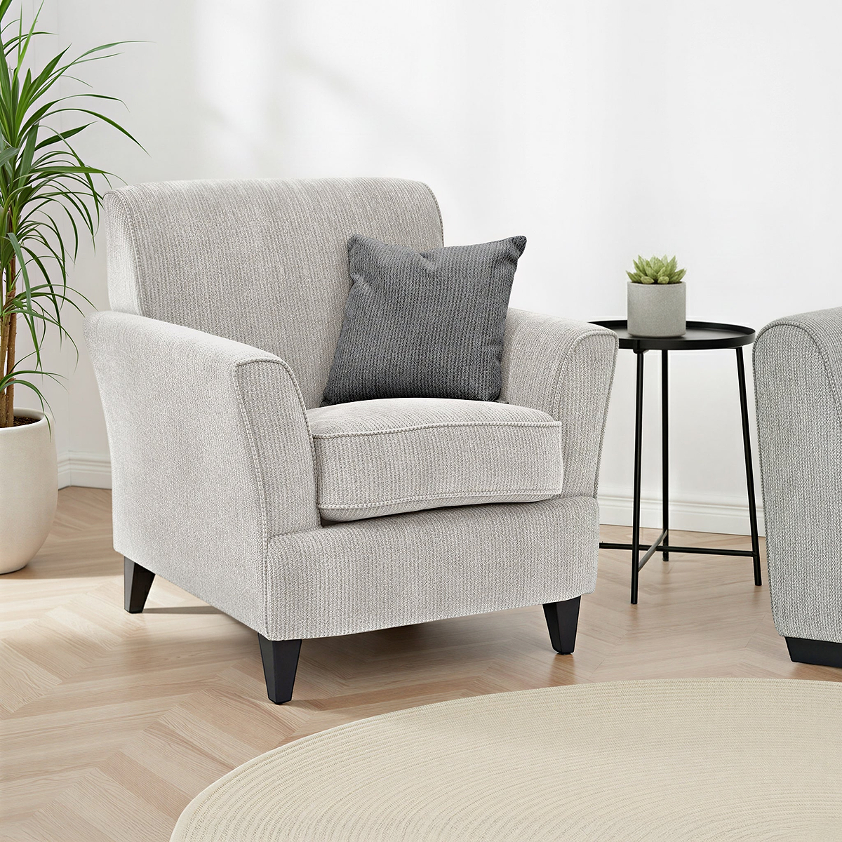 Thornby Accent Chair Charcoal Light Grey from Roseland Furniture in a living room or a lounge. the oom has white walls and light oak flooring. a cream colour rug covers the flooring and on either side of the chair are some plants. 