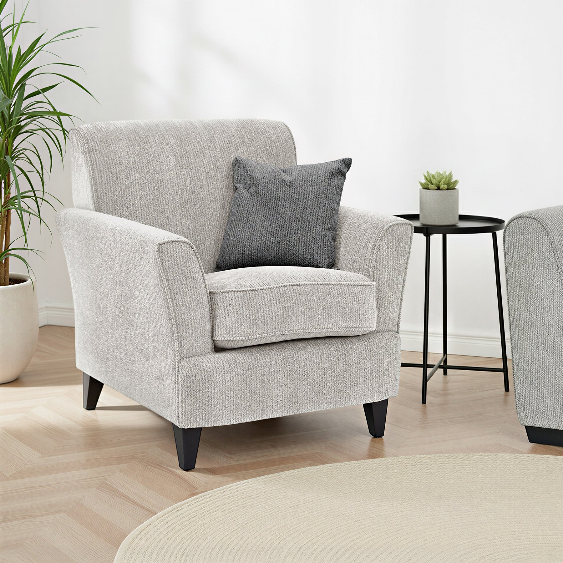 Thornby Accent Chair Charcoal Light Grey from Roseland Furniture in a living room or a lounge. the oom has white walls and light oak flooring. a cream colour rug covers the flooring and on either side of the chair are some plants. 