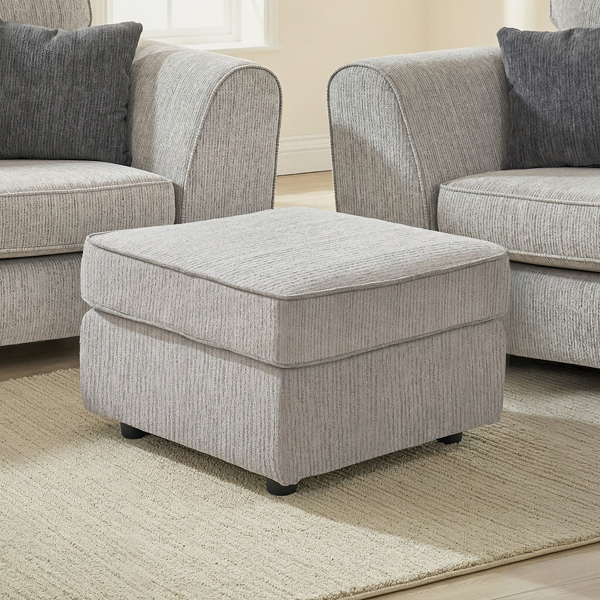 Thornby Standard Footstool Light Grey Charcoal from Roseland Furniture in a living room with the arm of a sof aon either sie, the room has white walls and light oak flooring covered by a cream rug.