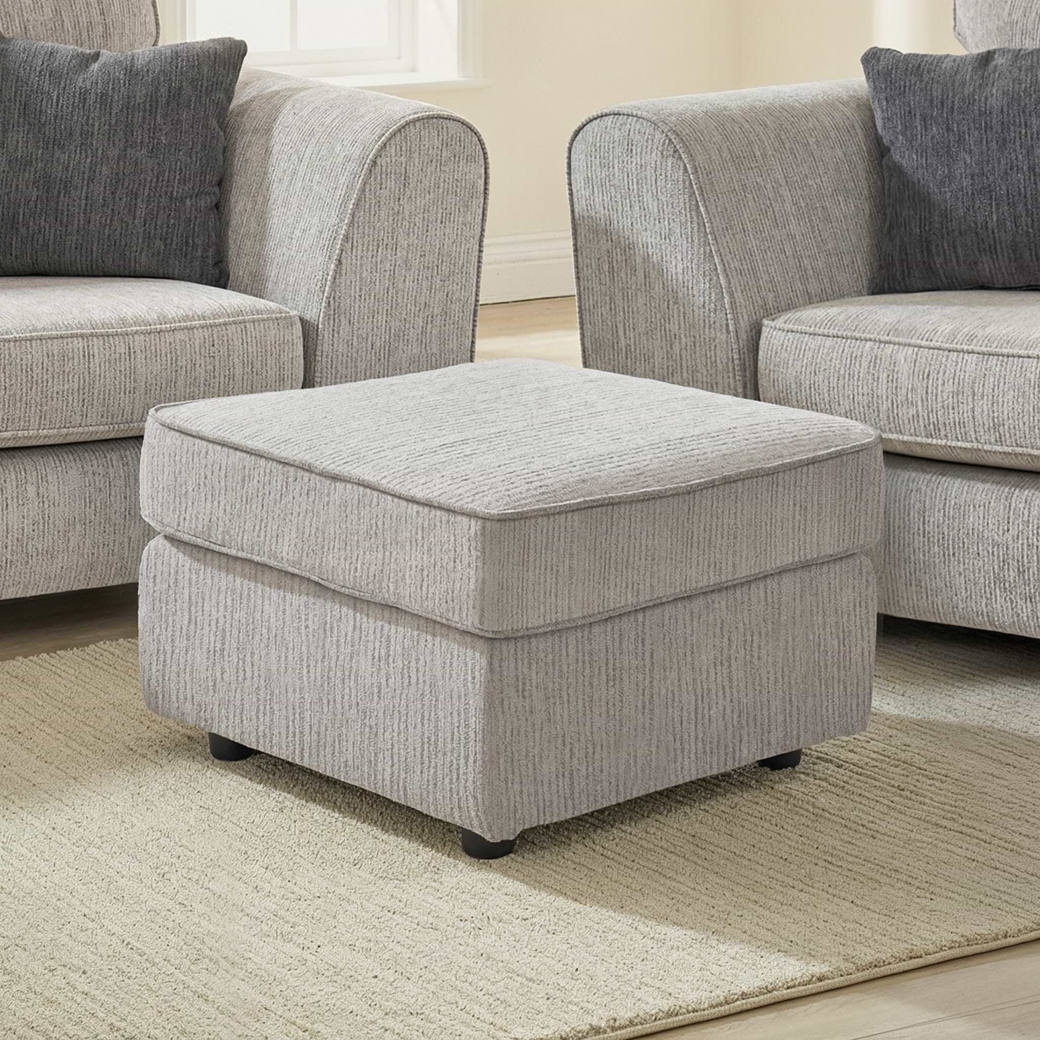 Thornby Standard Footstool Light Grey Charcoal from Roseland Furniture in a living room with the arm of a sof aon either sie, the room has white walls and light oak flooring covered by a cream rug.