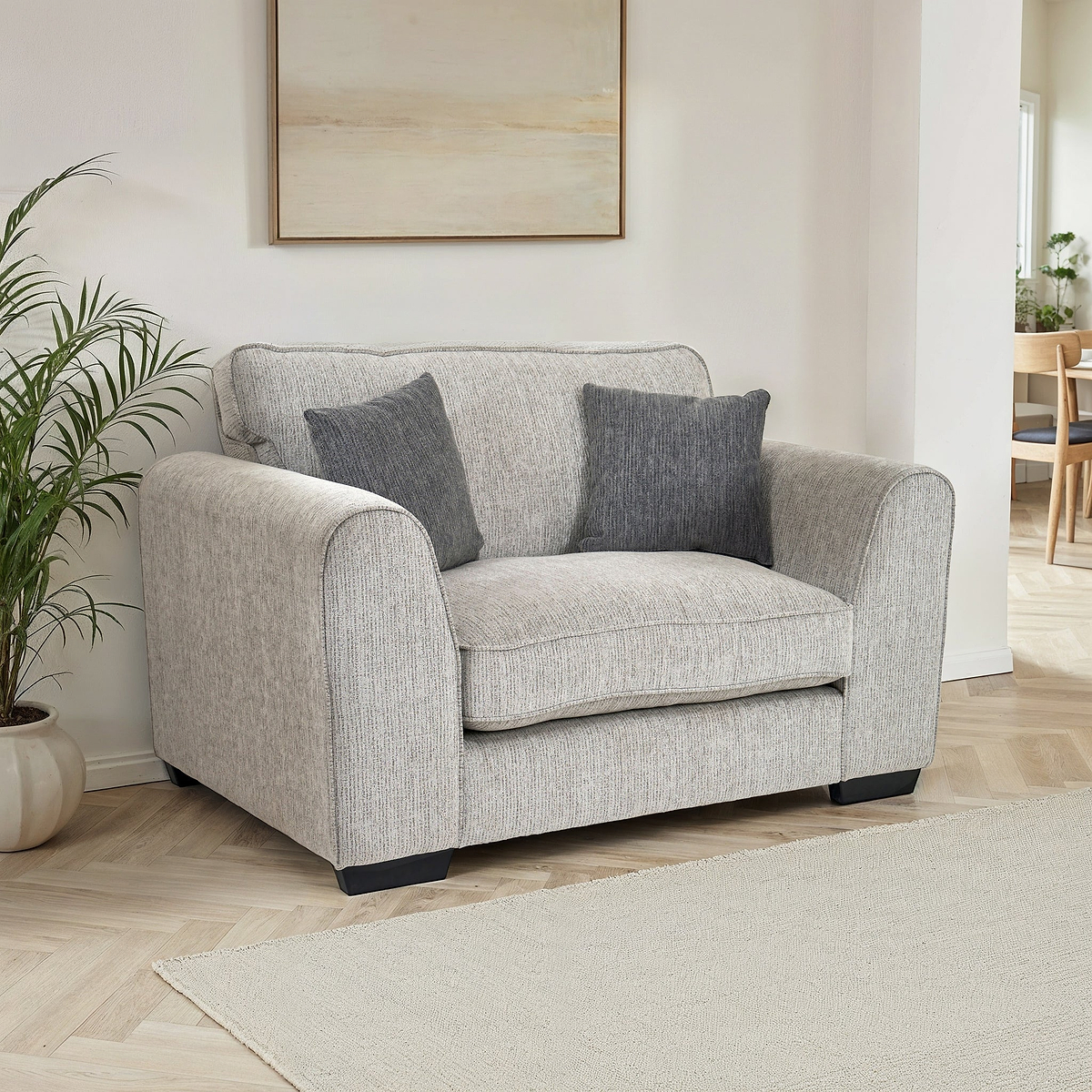 Thornby Snuggle Chair Light Grey Charcoal from Roseland Furniture positioned in a living room or lounge with white walls and light oak flooring, on the left is a plant on the right in the background is a dining room, above is a piece of abstract artwork and covering the floor is a light cream colour rug.
