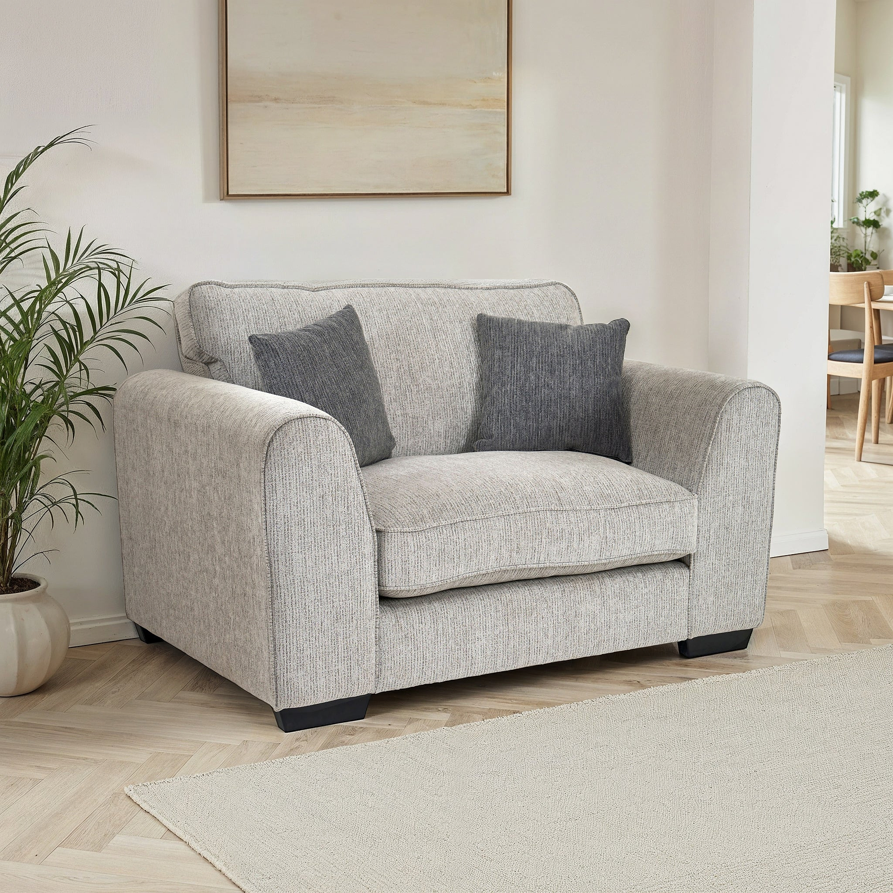 Thornby Snuggle Chair Light Grey Charcoal from Roseland Furniture positioned in a living room or lounge with white walls and light oak flooring, on the left is a plant on the right in the background is a dining room, above is a piece of abstract artwork and covering the floor is a light cream colour rug.