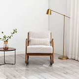 A beige armchair stands between a round side table with a vase and plant and a brass floor lamp, set against a white wall on a light wood floor.