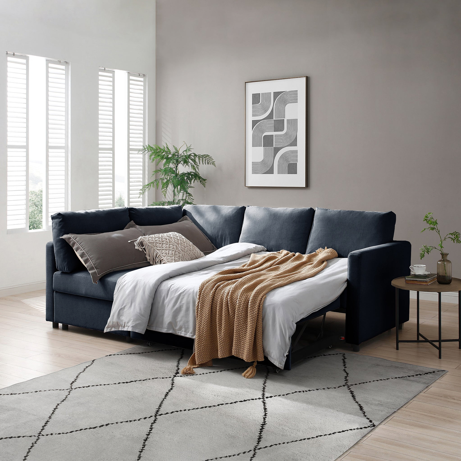 A navy-blue sectional sofa is adorned with pillows and a throw blanket in a minimalist living room, featuring a patterned rug, side table with plants, and framed artwork on the wall.