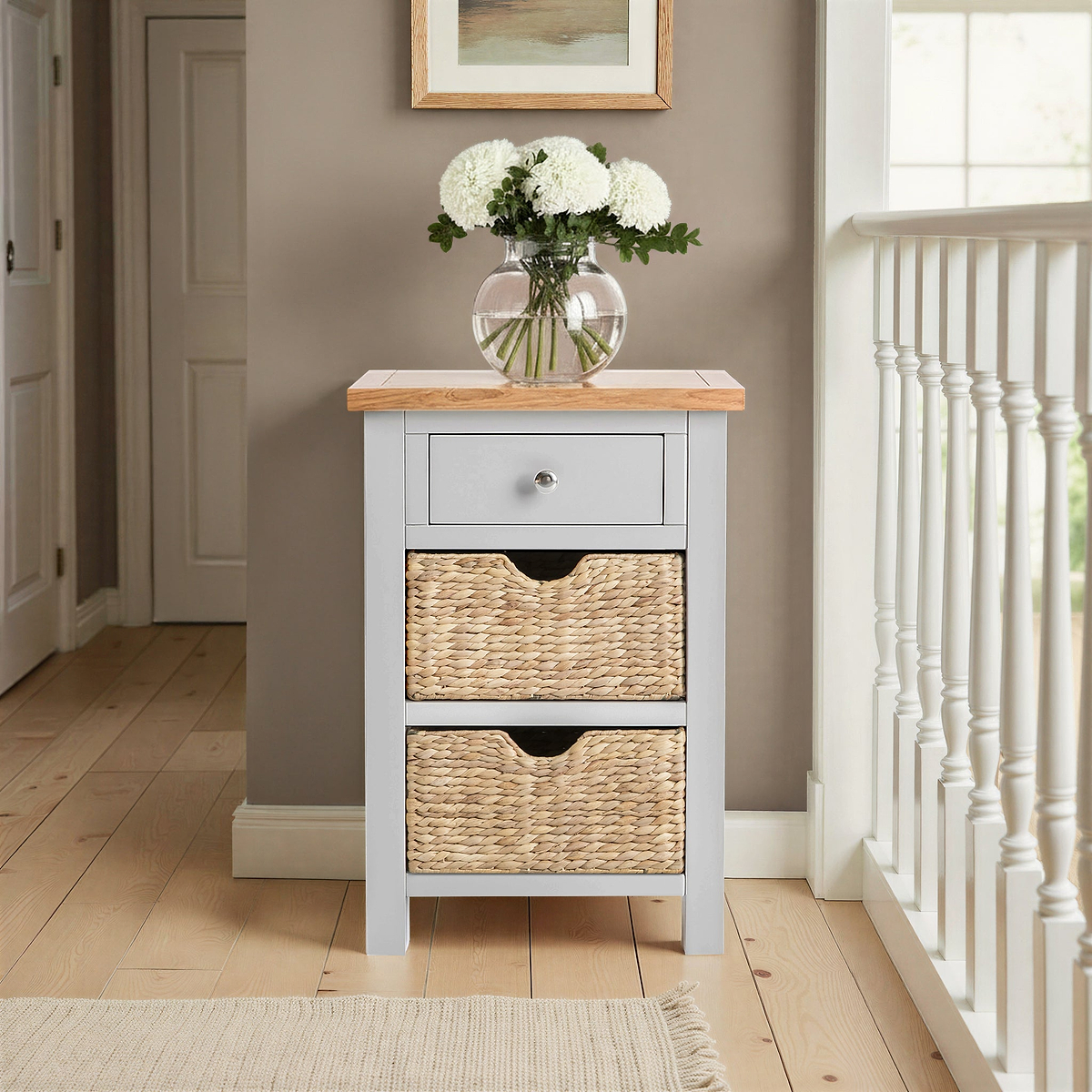 A small wooden storage cabinet with a drawer and two wicker baskets in a hallway with a vase of flowers on top