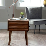 A wooden side table supports a metal lamp, books, and a potted succulent, situated near a window and a grey upholstered chair, in a well-lit room.