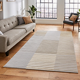 A striped area rug lies on a wooden floor, flanked by a grey sofa and a black armchair, with large windows revealing a cityscape.