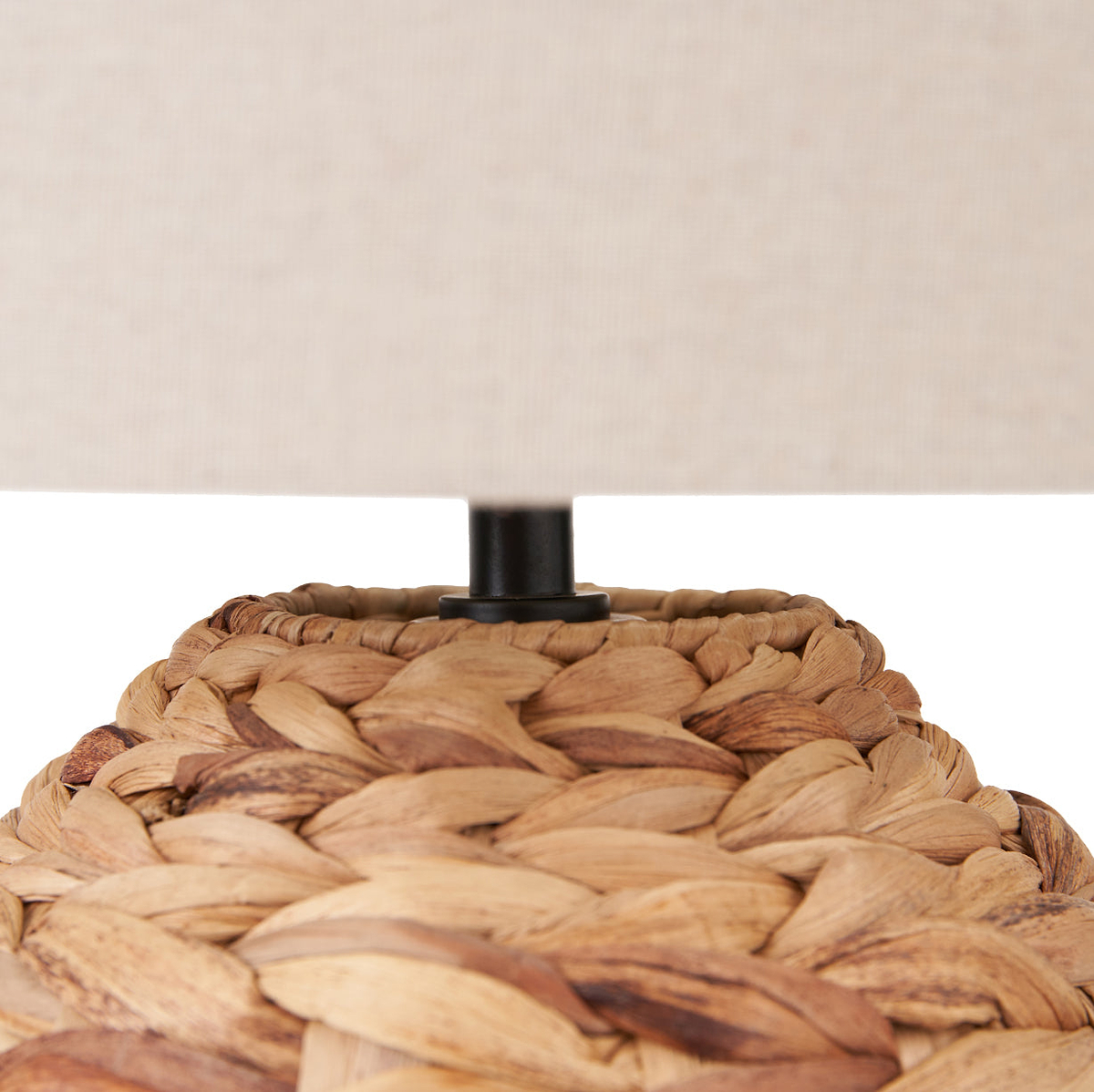 Close-up of a woven natural fiber lamp base with a black central stem and a beige fabric lampshade partially visible against a white background.