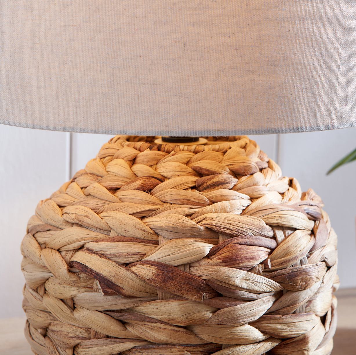 A close-up of a woven natural fiber lamp base with a beige fabric drum shade, shown against a light neutral background.