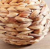 A close-up of a round woven basket made from natural water hyacinth fibers, showing the textured interlaced weave on a light wood surface.